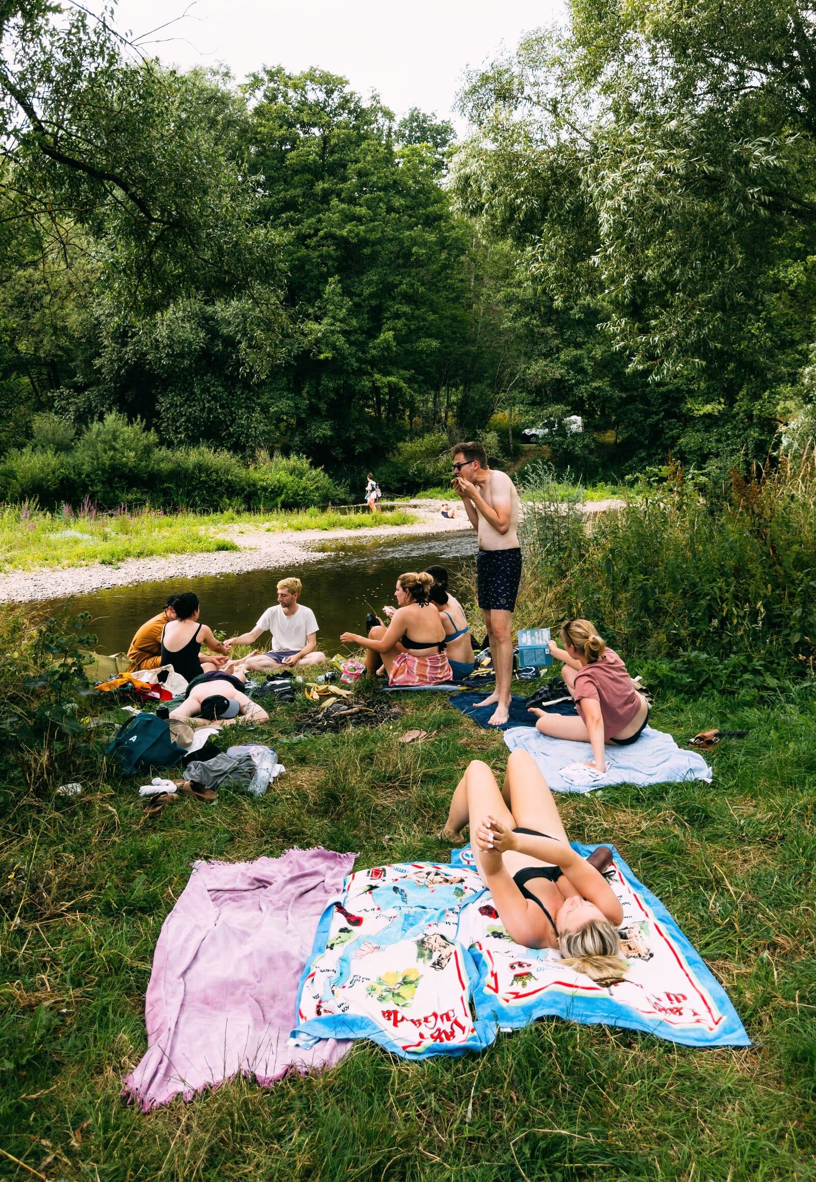 The friend group relaxing on towels by a river on a summer day